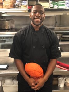 An alumni wears a black chef's coat in the kitchen of a restaurant in DC.