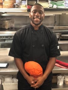 An alumni wears a black chef's coat in the kitchen of a restaurant in DC.