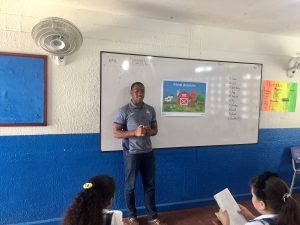 A student in a gray shirt and blue pants stands in front of a whiteboard in a classroom in Colombia.