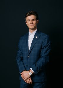 A formal portrait of Omar Alshogre features the Georgetown student standing with his hands clasped wearing a navy blue shirt and light blue button-up.