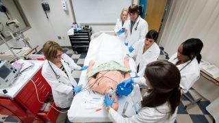 Students and professors in white coats around a fake body