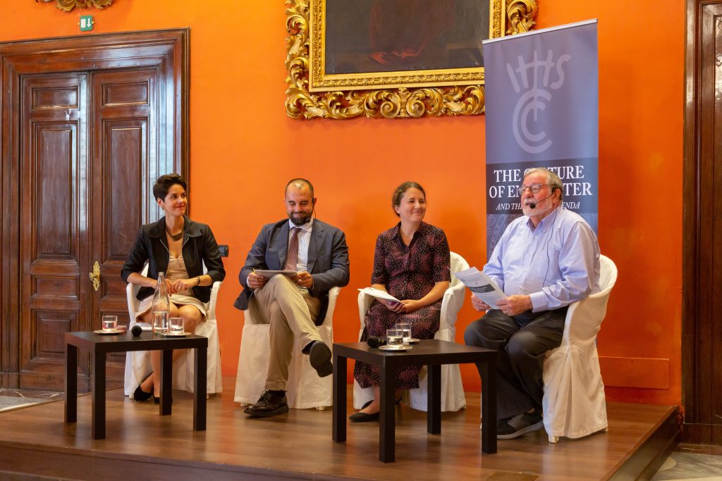 Four conference panelists sit on a stage before an orange wall at a conference in Rome.