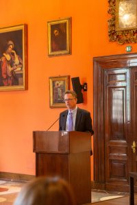 Tom Banchoff, vice president for global engagement, stands behind a podium and delivers a speech in front of an orange wall with framed pictures.