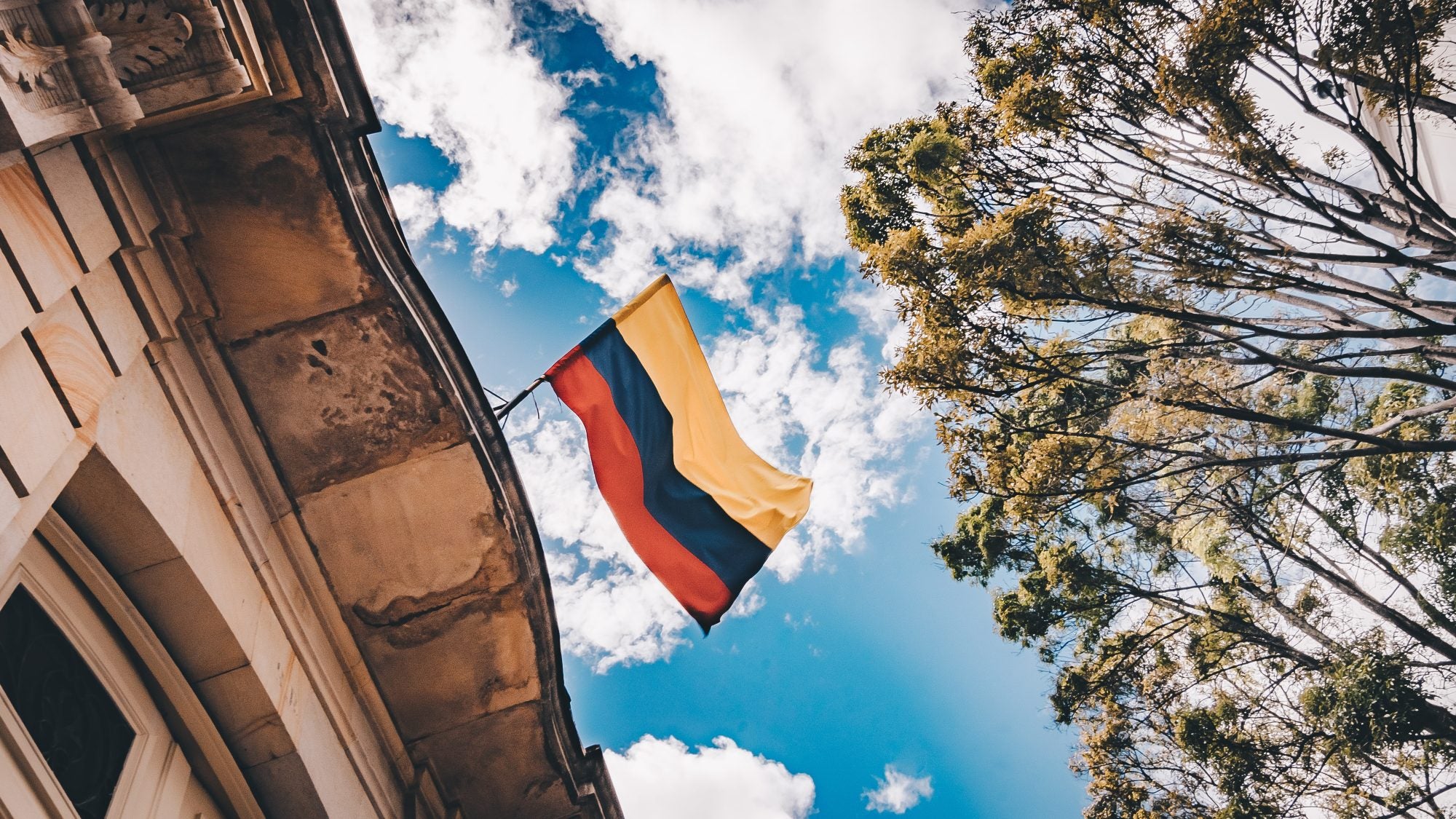 Colombian flag flying above building with blue sky and a tree above it.