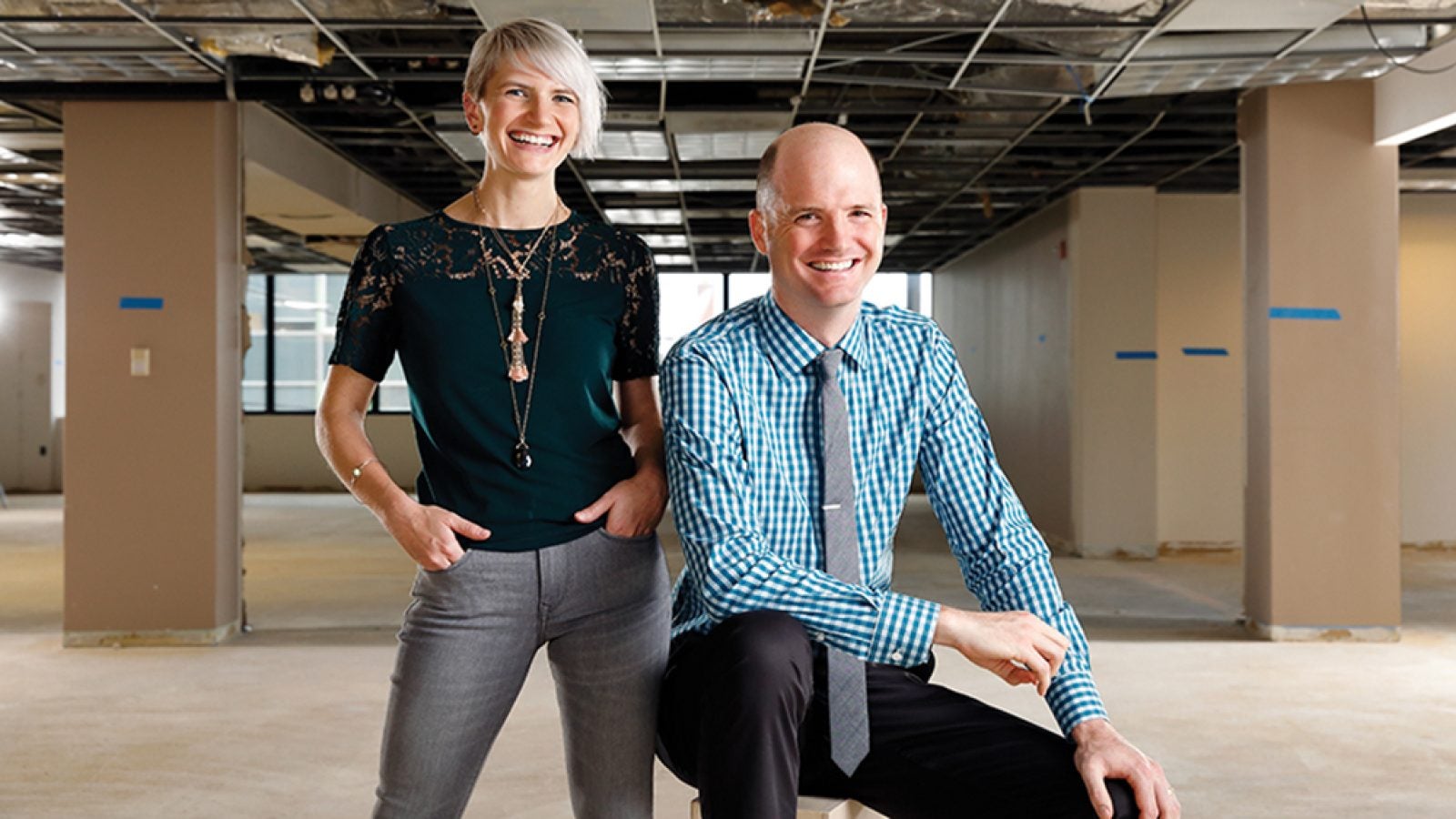 One alumni, dressed in a black shirt and jeans, stands next to another business school alumni, who&#039;s dressed in a blue shirt, a tie and black pants.