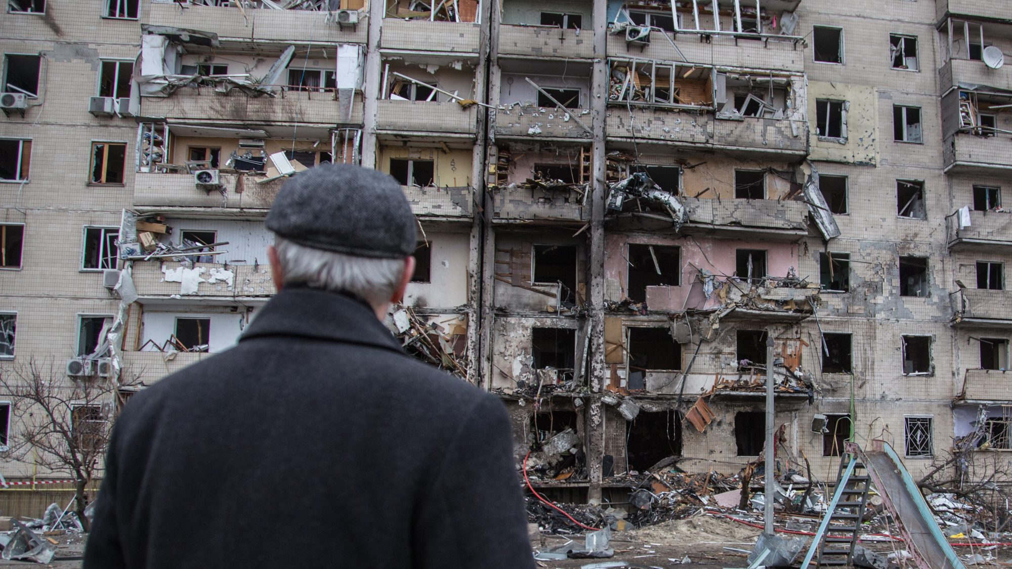 A man in a coat and hat looks at a partially destroyed apartment building in Kyiv