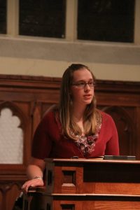 A woman with glasses wears a read short-sleeve cardigan and speaks from behind a podium in a church