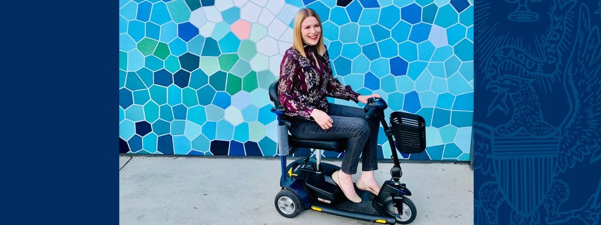 A white woman with straight, blonde hair, wearing a burgundy patterned shirt and grey jeans sits on a blue and black mobility scooter and smiles in front of a wall with a variety of blue and white geometric shapes