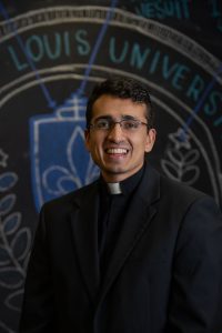 Headshot of Christian Verghese, S.J. wearing a black blazer and priest's collar in front of formal seal background