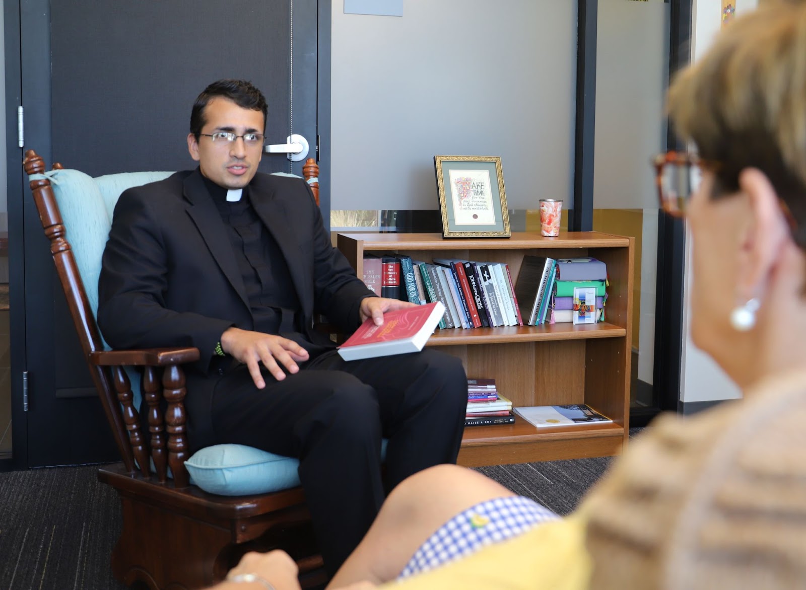 A woman with glasses looks at Christian Verghese who holds a book and wears a priest's collar in a classroom