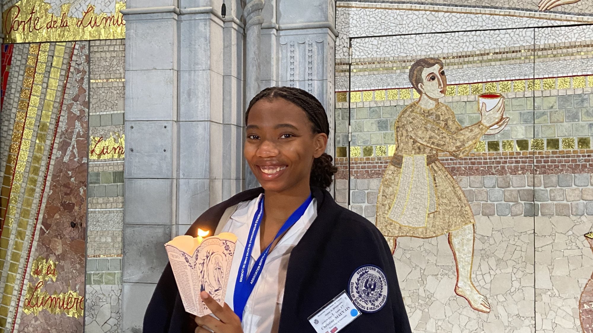 Christine Mauvais wears a white dress, blue medal and black coat off her shoulders while holding a candle in front of mosaic religious art