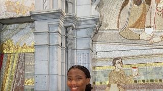 Christine Mauvais wears a white dress, blue medal and black coat off her shoulders while holding a candle in front of mosaic religious art