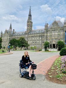 A white woman with straight, blonde hair, wearing a navy dress and a black blazer sits on a blue and black mobility scooter and smiles in front of Healy Hall with a blue sky behind.