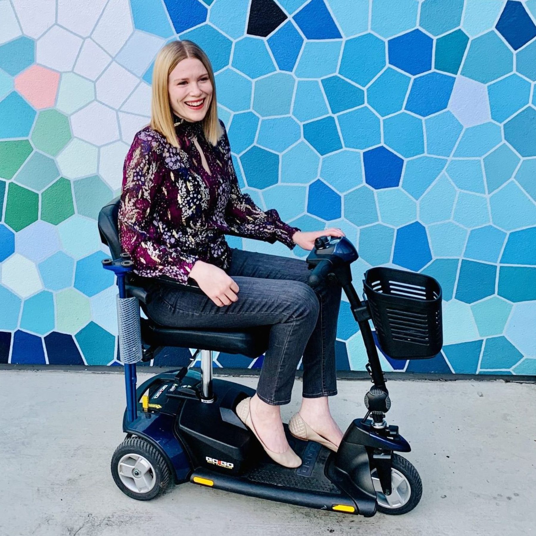 A white woman with straight, blonde hair, wearing a burgundy patterned shirt and grey jeans sits on a blue and black mobility scooter and smiles in front of a wall with a variety of blue and white geometric shapes.