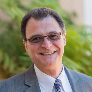 A man wearing a suit with gelled-back hear and glasses smiles at the camera in front of trees