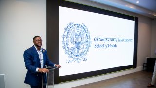 Christopher King at a clear podium in front of a projection with the text &quot;Georgetown University School of Health&quot;