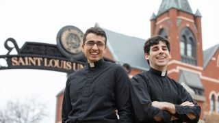 Two young men wearing priest&#039;s collars in front of a church
