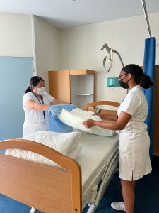 Two nursing students stand over a nursing bed in a hospital and put a blue pillowcase on a white pillow.