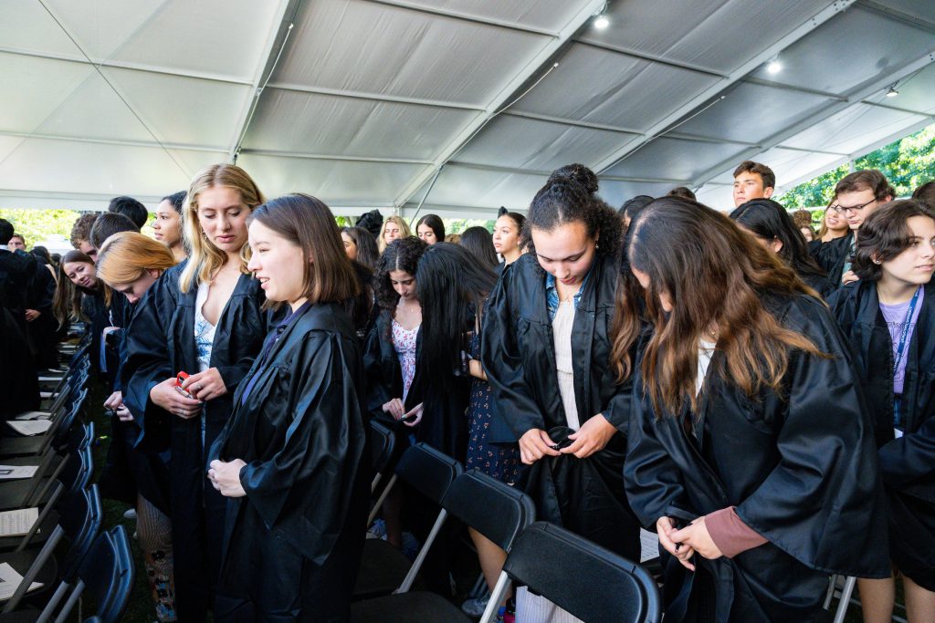 New students sitting in folding chairs under a tent outdoors put on black academic robes.