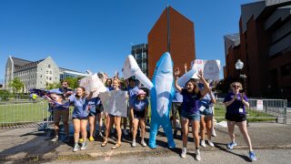 Students in matching T-shirts and one dressed as a shark hold signs and cheer