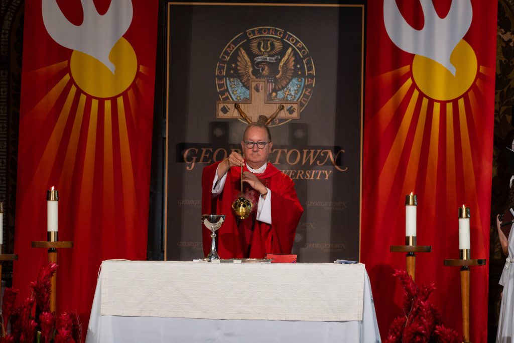 A priest in red robes moves incense over a table in front of a Georgetown sign in between two red banners