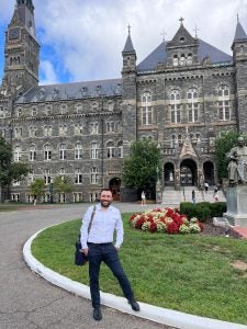 Man in light blue button-down shirt and dark pants stands in front of Healy Hall and the John Carroll statue