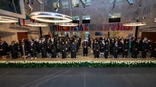 Students wearing academic robes stand in a room with dozens of hanging flags