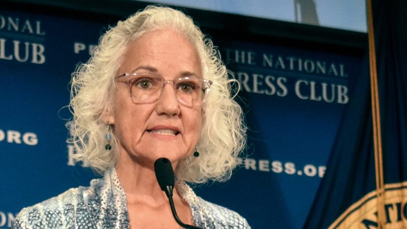 Woman with white hair and glasses speaks into microphone from behind a podium