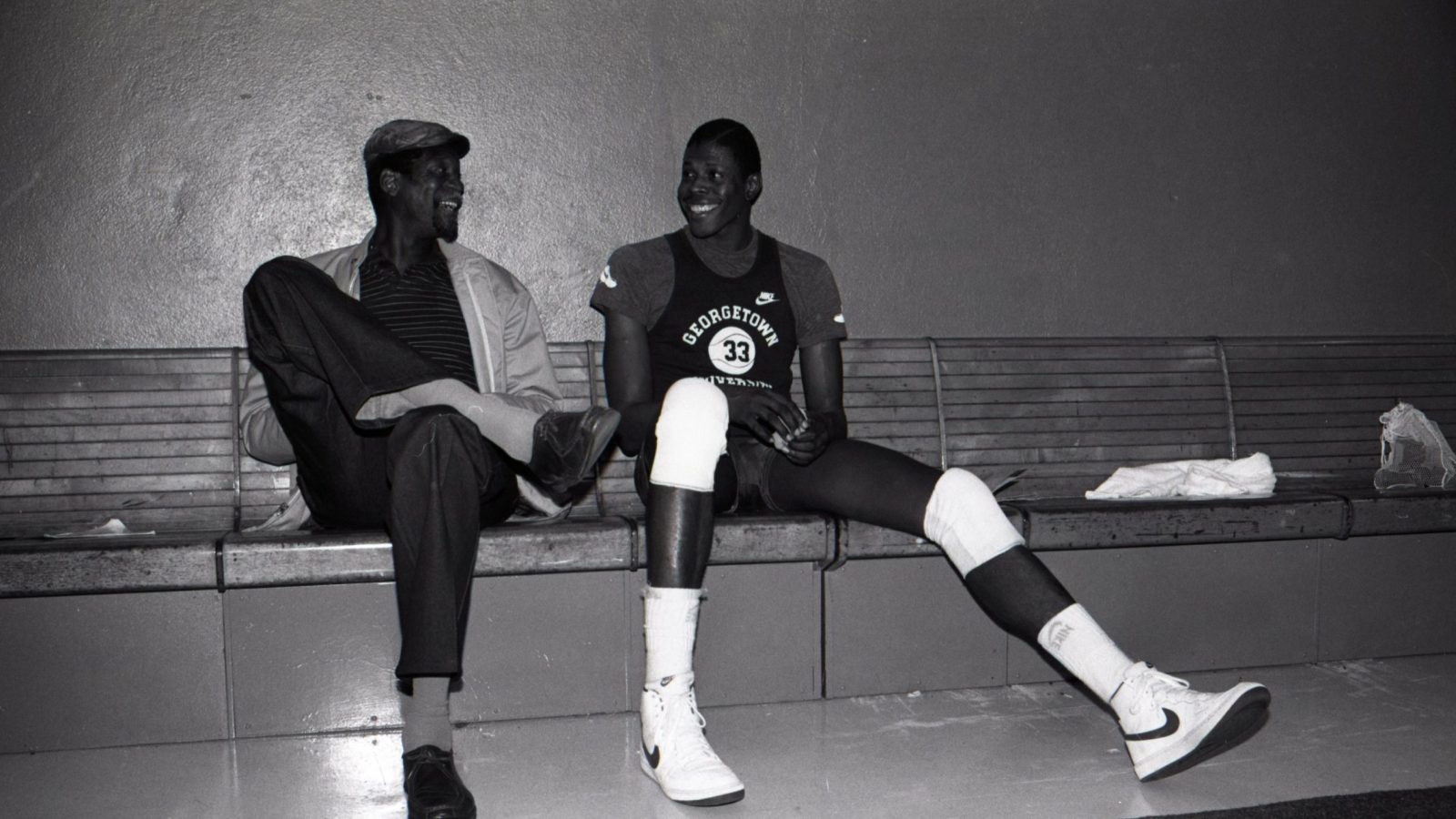 Black and white image of Russell (left) wearing a hat sitting on a bench next to Patrick Ewing (right) in a Georgetown basketball uniform
