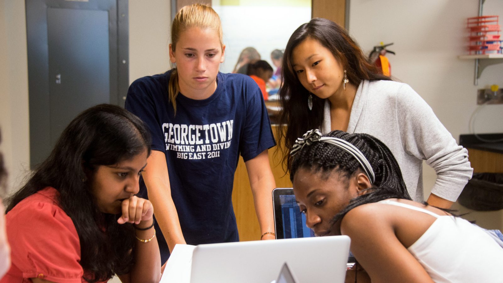 A group of four students look at a laptop screen together in a classroom setting.