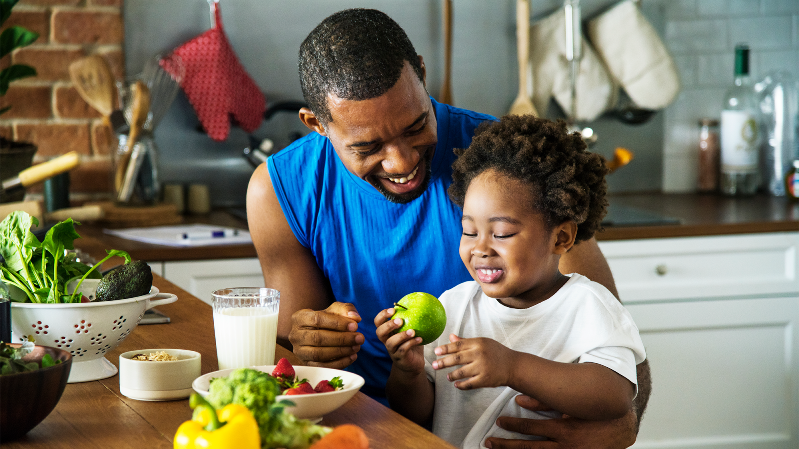 A father feeds his child fresh fruit as they both sit at the kitchen table.
