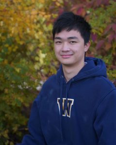 A young man wears a blue hoodie in front of trees with changing leaves