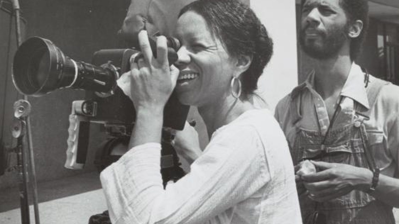 B&amp;W photo of Kathleen Collins, a Black woman with hair tied behind a camera, one eye pressed against the objective, she is wearing a white long sleeved shirt, a Black man is standing in the background