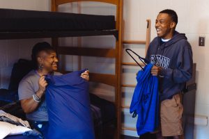 Briana Green sits on her son's bed in his dorm room and puts a blue pillow case on his pillow. Keiran Green, who is wearing a Georgetown sweatshirt, stands next to her holding a hanger and laughing during move-in 2022.