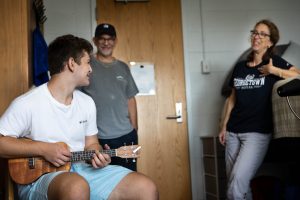 Brian Kaye (SOH’26) sits on a chair in his dorm room and plays the ukulele. He looks over at his parents, who are standing behind him and smiling.