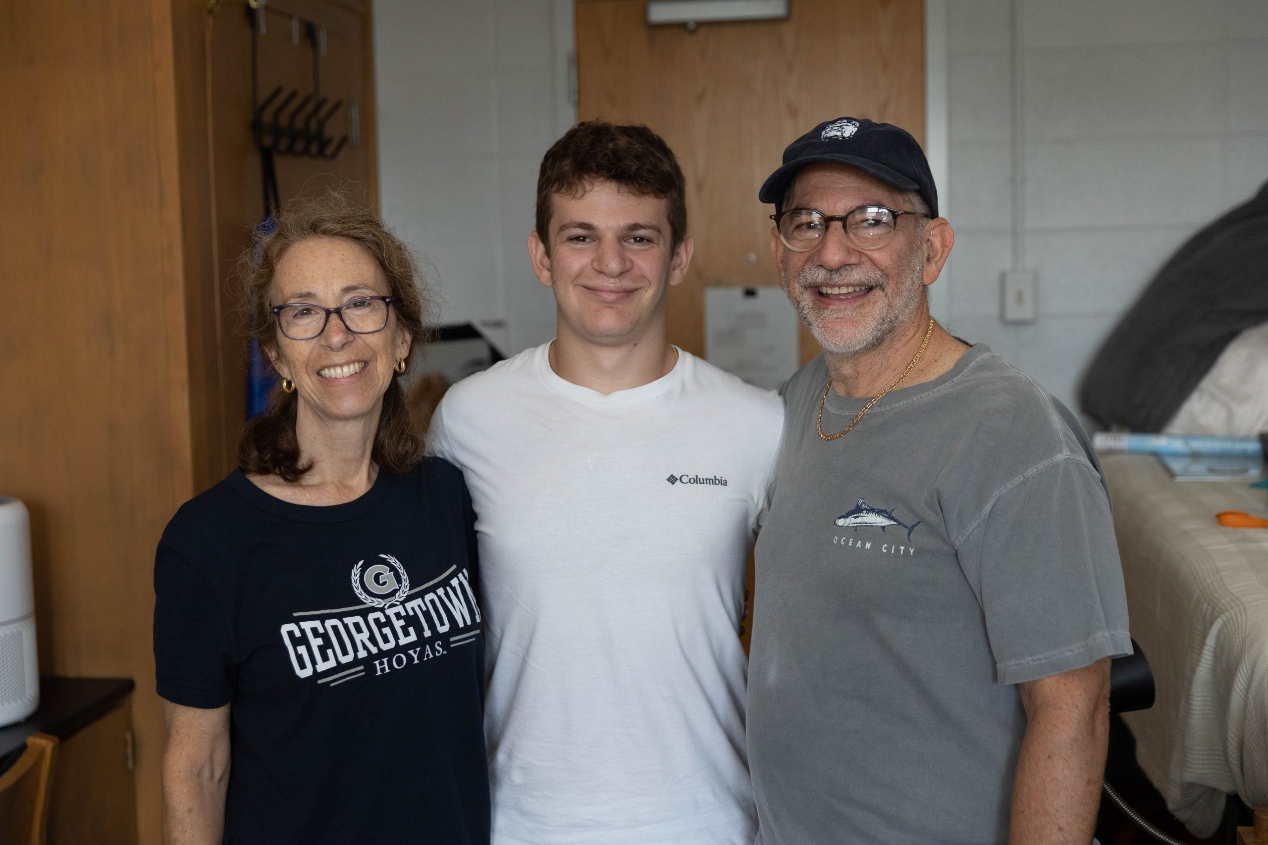 Brian Kaye (SOH’26) stands in between his two parents in his dorm room at Georgetown. They are all smiling at the camera.