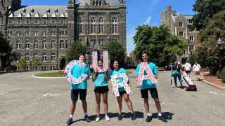 Four students in light blue T-shirts hold letters spelling out CURA while standing in front of Healy Hall