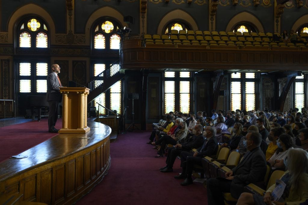 A man in a suit speaks from behind a podium on a stage addressing students wearing masks in the audience. 