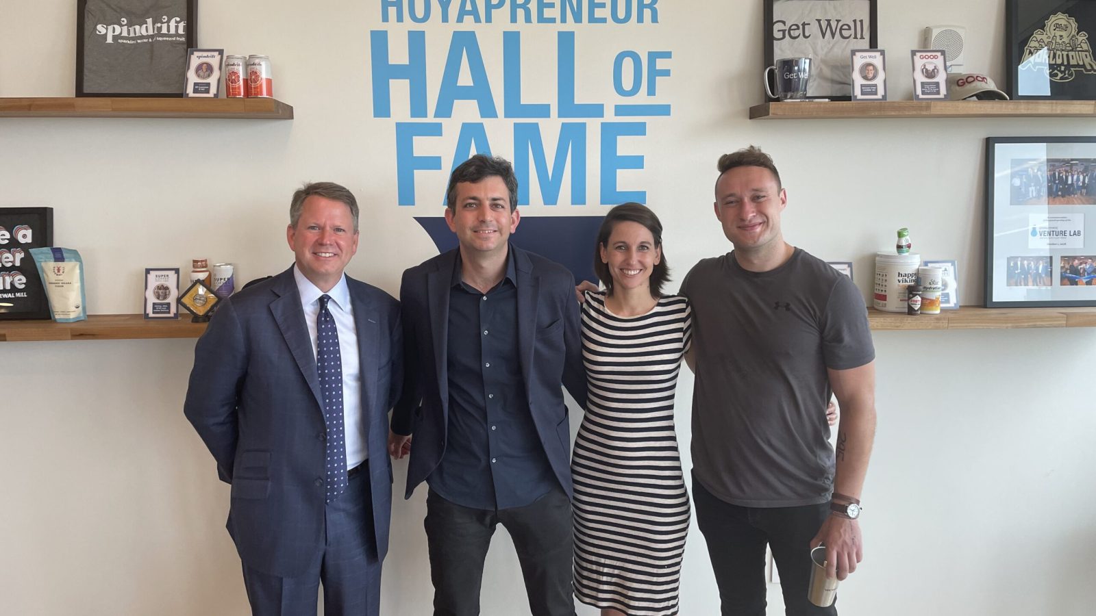 Four Georgetown community members stand in front of a sign painted on a wall that says &quot;Hoyapreneur Hall of Fame&quot;