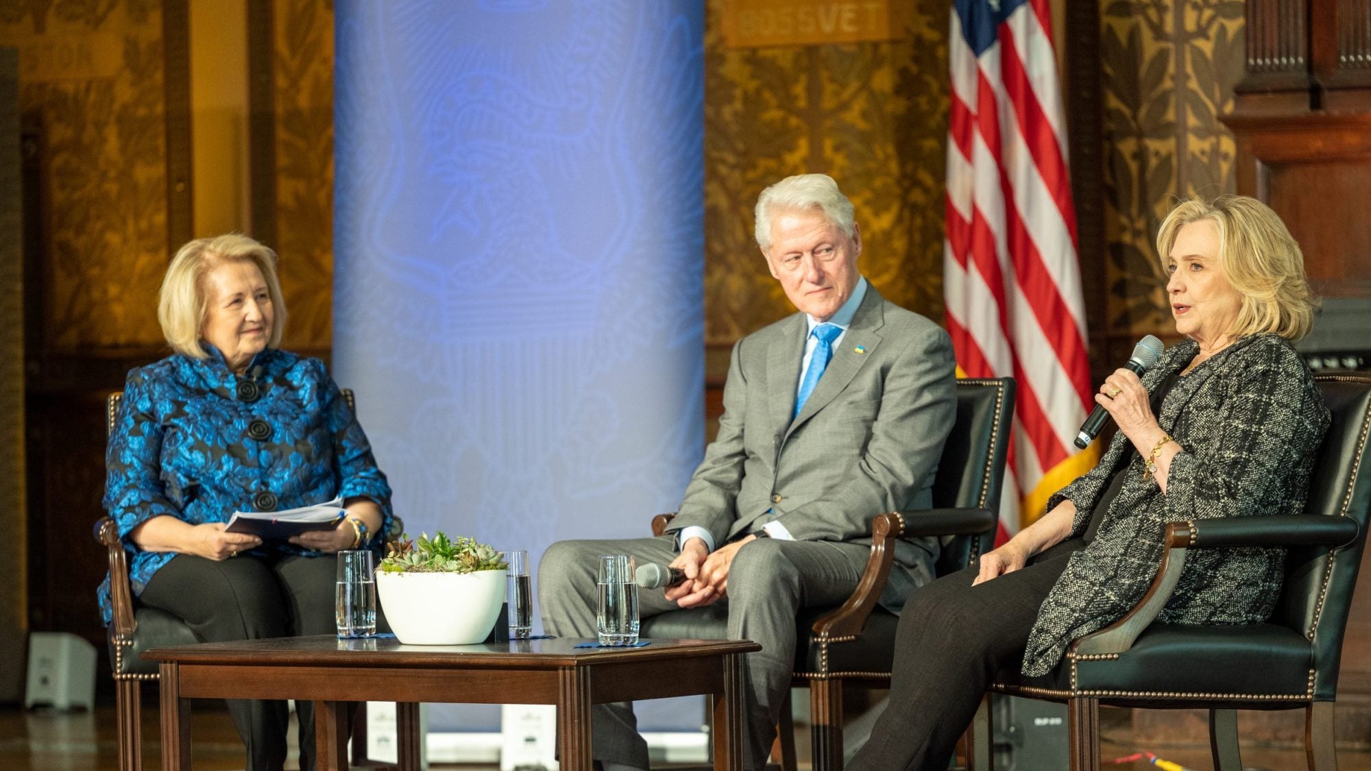 Melanne Verveer and Bill and Hillary Clinton sit on stage while Hillary speaks into a microphone