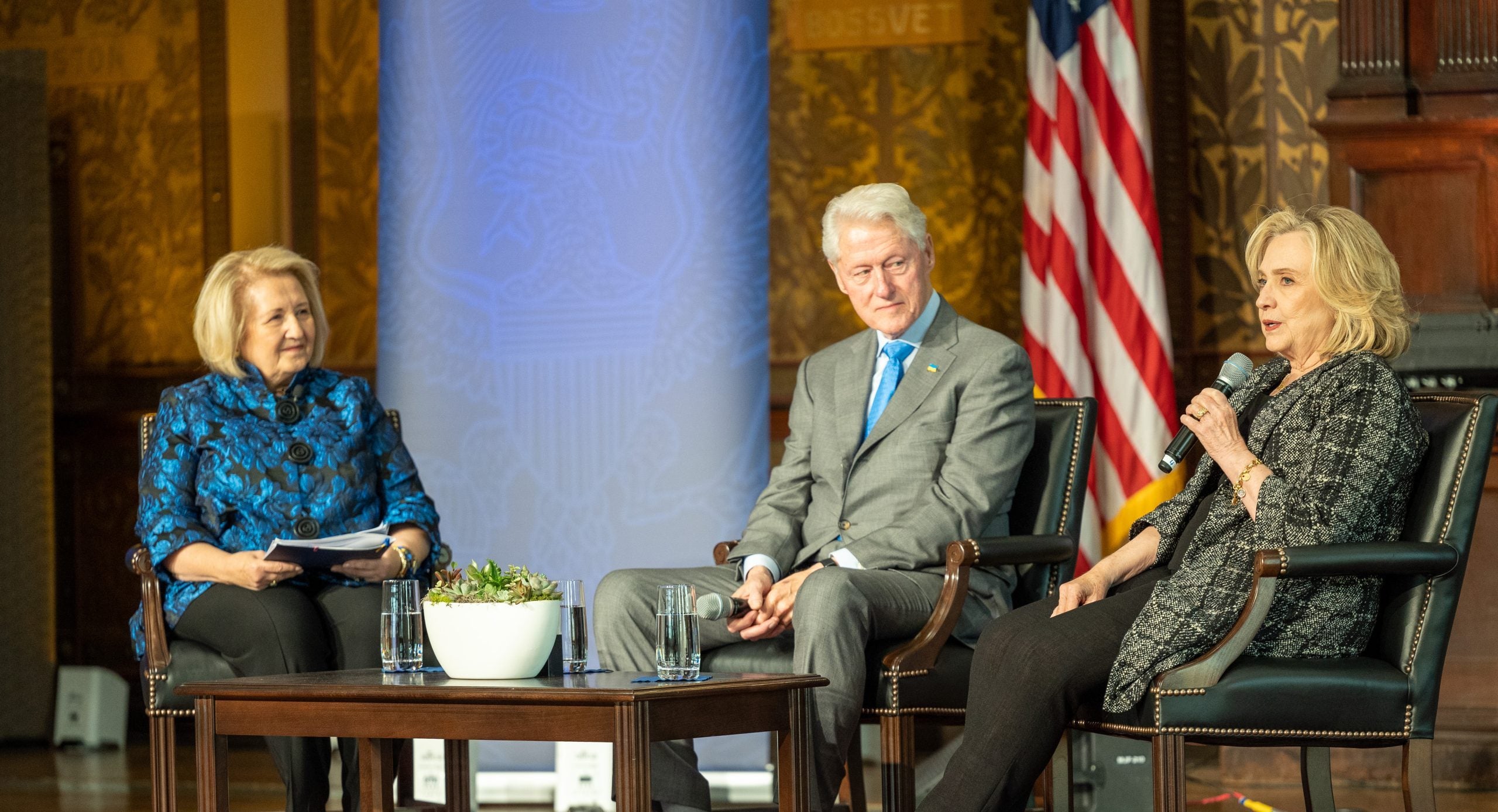 Melanne Verveer and Bill and Hillary Clinton sit on stage while Hillary speaks into a microphone