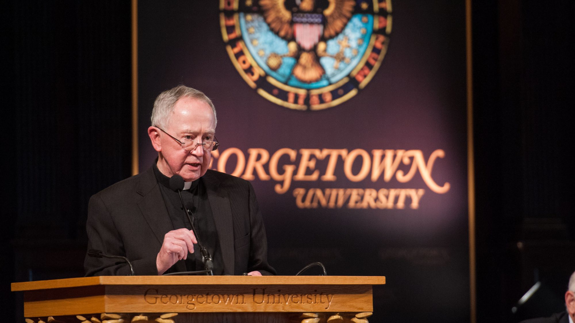Fr. O&#039;Malley wears glasses, a priest&#039;s collar and black clothes while giving a speech in front of a Georgetown University sign