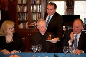 Fr. O'Malley makes a suprised face at a piece of cake with a candle while President DeGioia (right) laughs