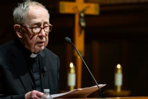 Fr. O'Malley speaks into a podium microphone with a crucifix and candles in the background