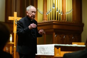 Fr. O'Malley speaks with glasses in his hand in front of a crucifix and pipe organ in a church