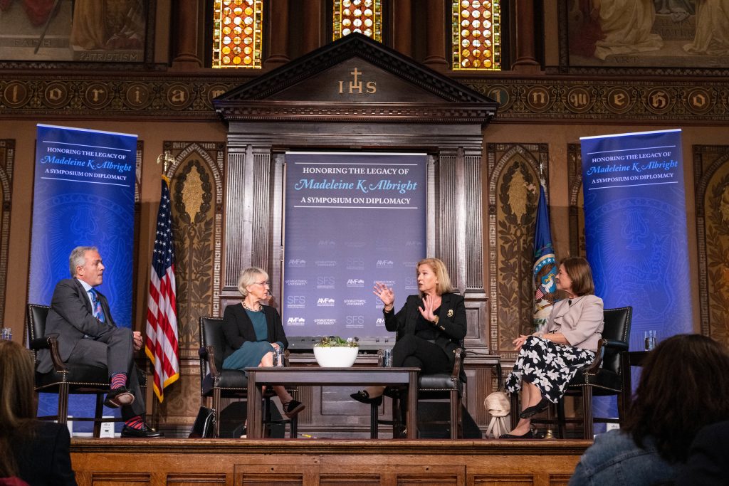 Four panelists having a discussion on Gaston Hall's stage with signs reading "Honoring the Legacy of Madeleine K. Albright"