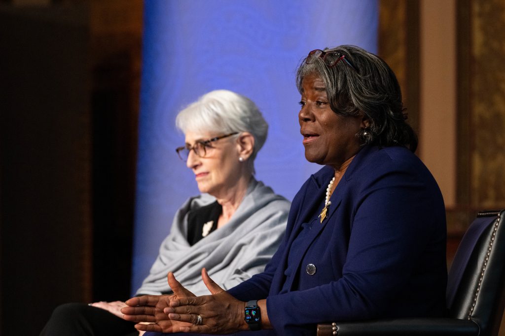 Wendy Sherman wears a gray wrap and glasses and Linda Thomas-Greenfield wears a blue pansuit while they sit onstage for a panel.