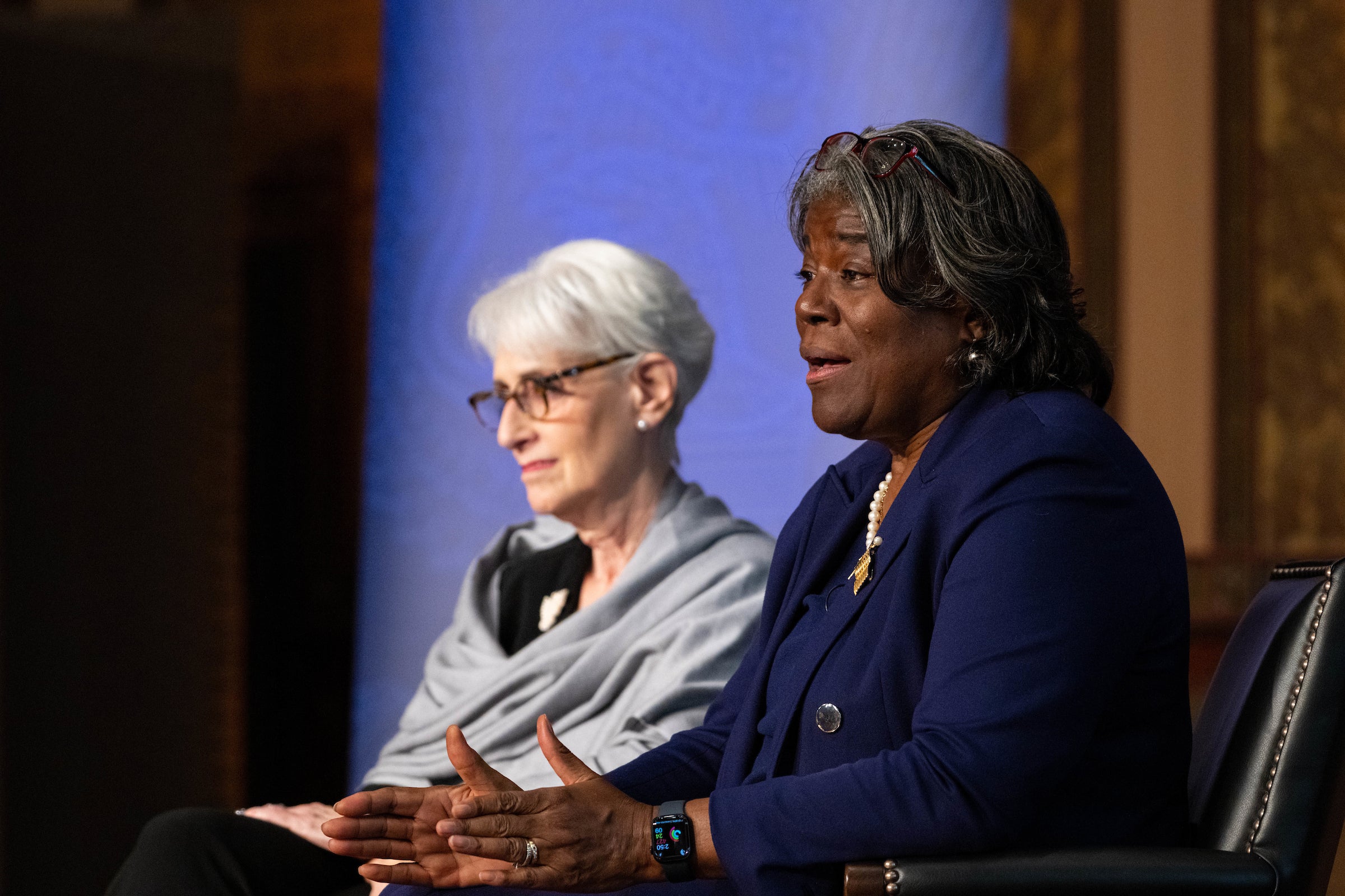 Wendy Sherman wears a gray wrap and glasses and Linda Thomas-Greenfield wears a blue pansuit while they sit onstage for a panel.