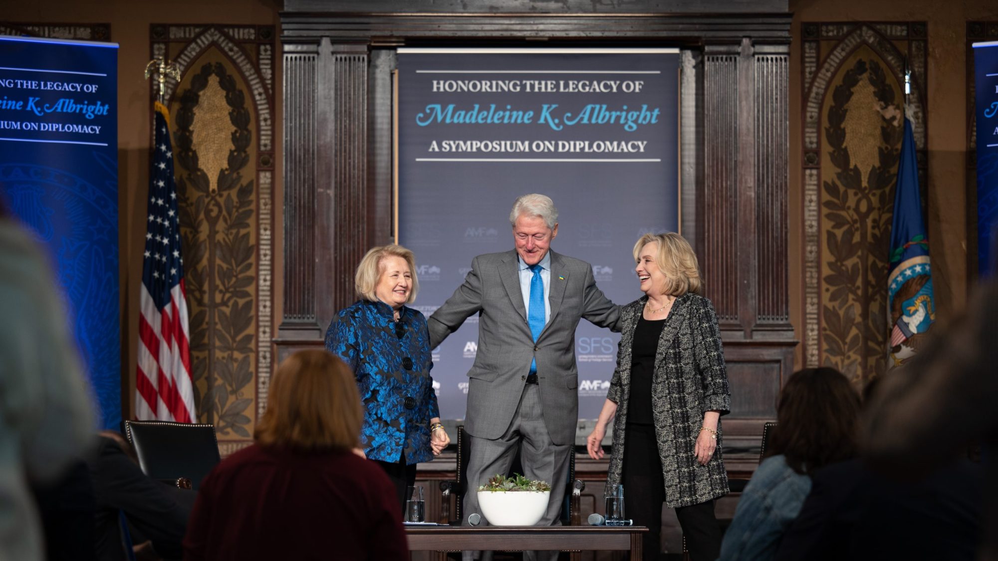 Melanne Verveer, Bill Clinton and Hillary Clinton stand on a stage with a banner behind them with the text &quot;Honoring the Legacy of Madeleine Albright&quot;