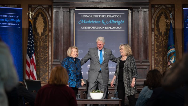 Melanne Verveer, Bill Clinton and Hillary Clinton stand on a stage with a banner behind them with the text 
